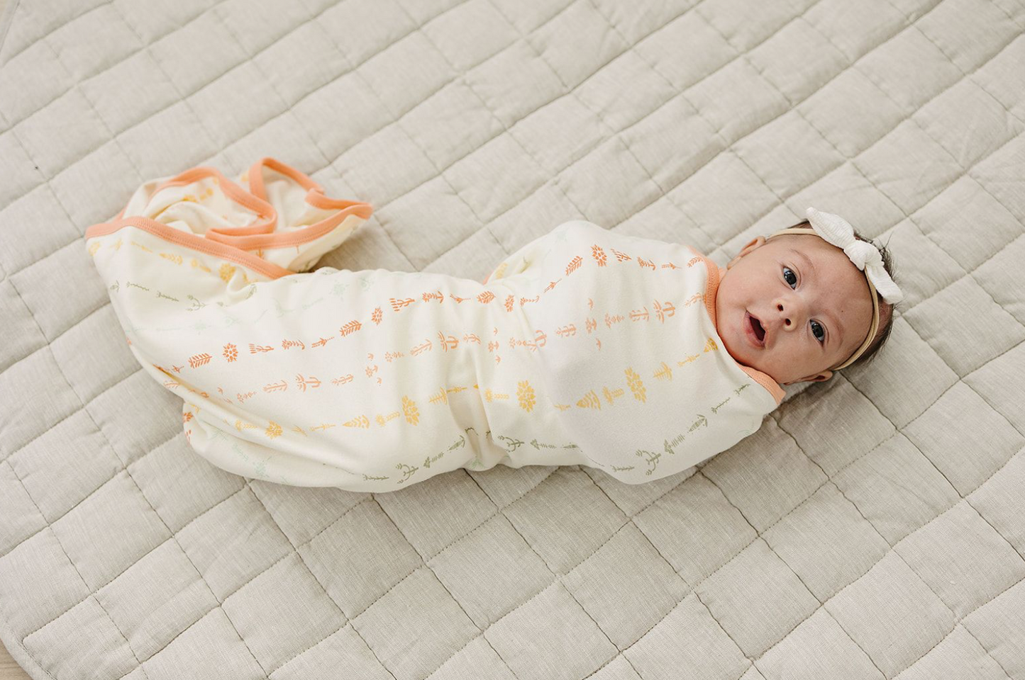 Baby swaddled in a white blanket with orange patterns on a beige quilted surface