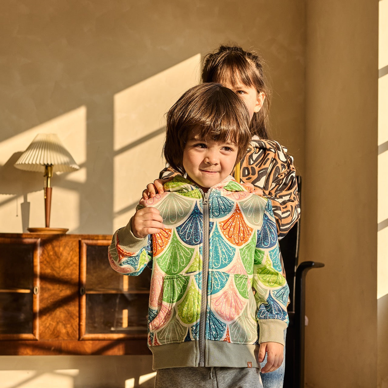Two children wearing colorful jackets in a warm, sunlit room.