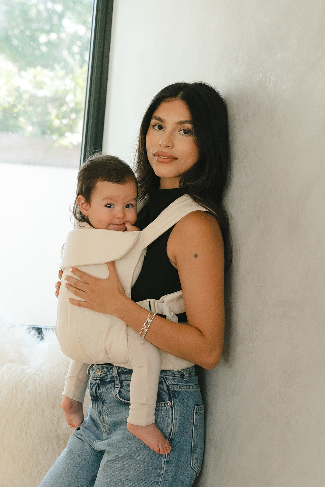 Woman holding a baby in a carrier against a light-colored wall.