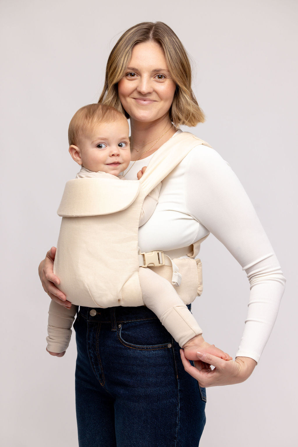Woman holding a baby in a beige baby carrier against a plain background