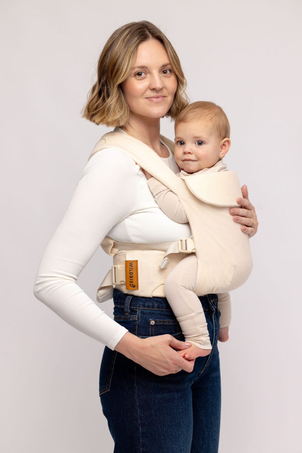 Woman holding a baby in a beige baby carrier against a plain background