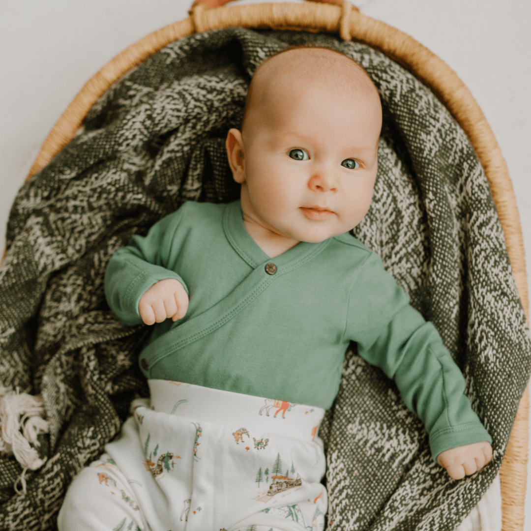 Baby in a green outfit sitting in a woven basket with a textured blanket