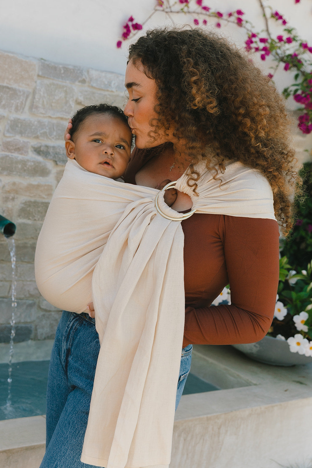 Woman holding a child in a sling outdoors with flowers in the background