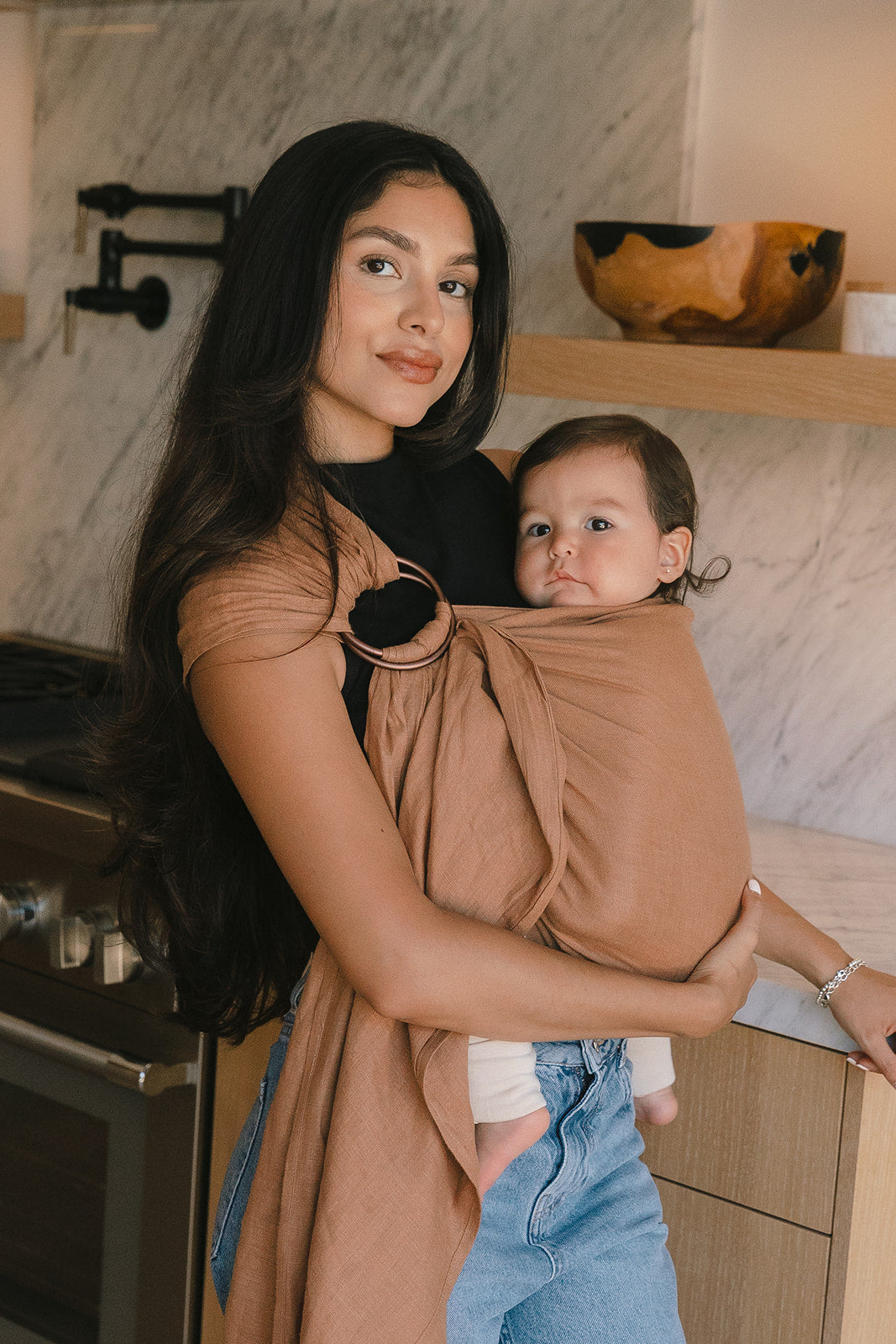 Woman holding a baby in a sling inside a kitchen.