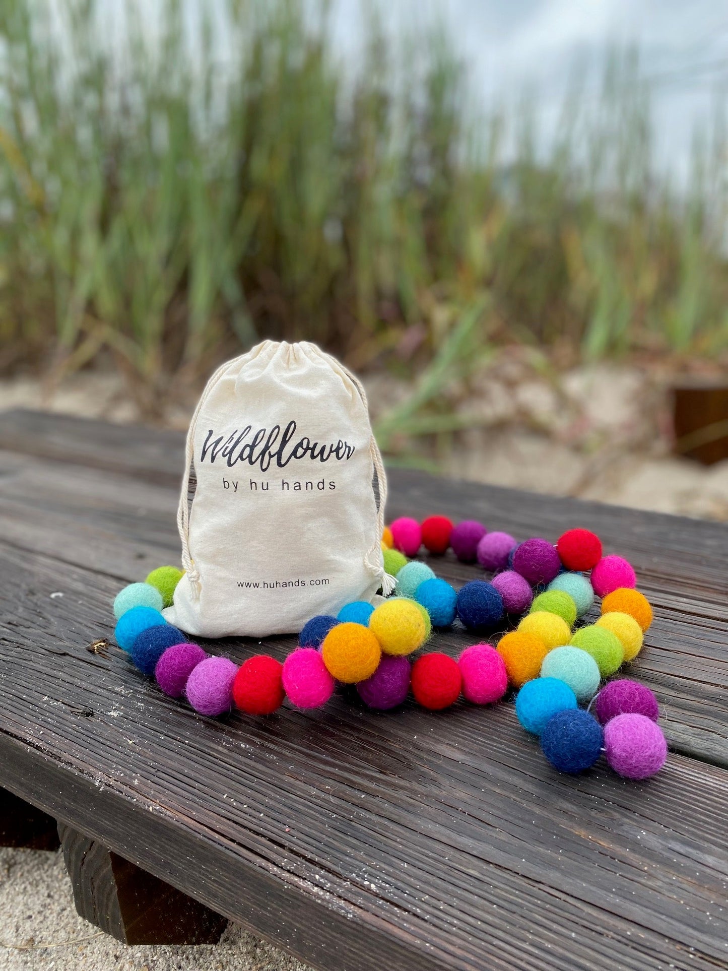 Colorful pom-pom bracelet on a wooden surface with a branded bag in the background.