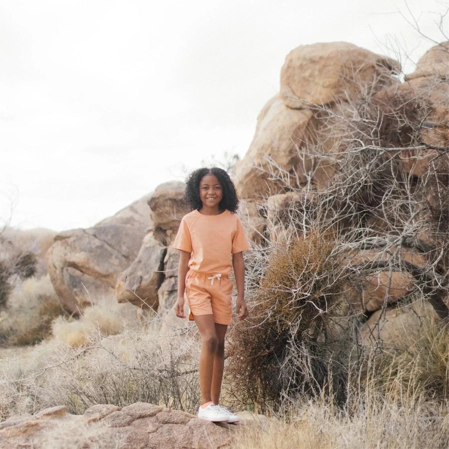 Person in an orange outfit standing in a desert landscape with large rocks and sparse vegetation.