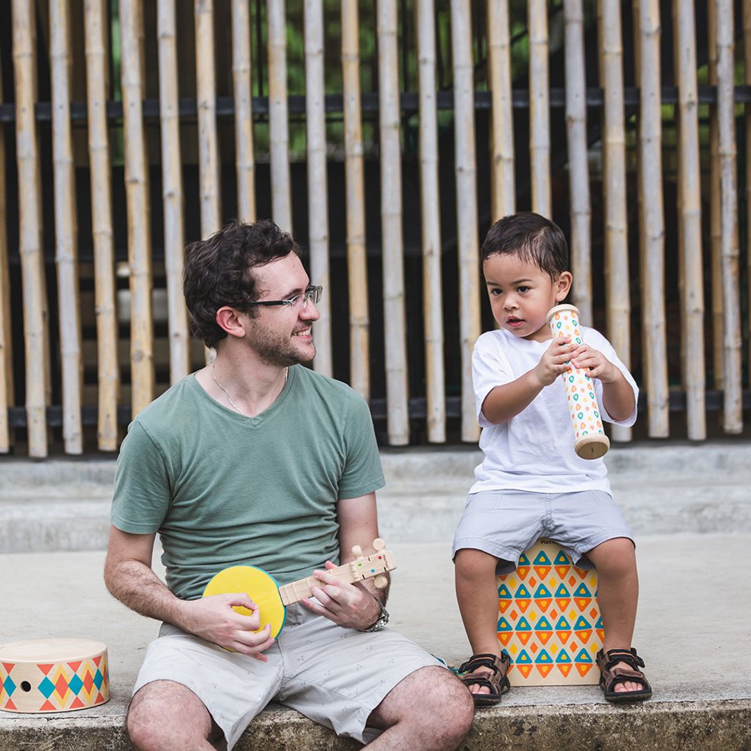 Man and child playing musical instruments outdoors with a bamboo fence in the background