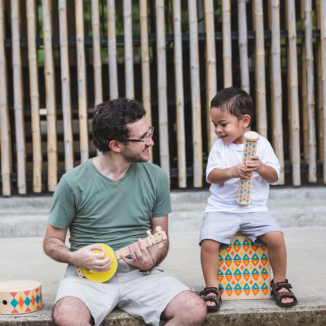 Man and child playing musical instruments outdoors with a bamboo fence in the background