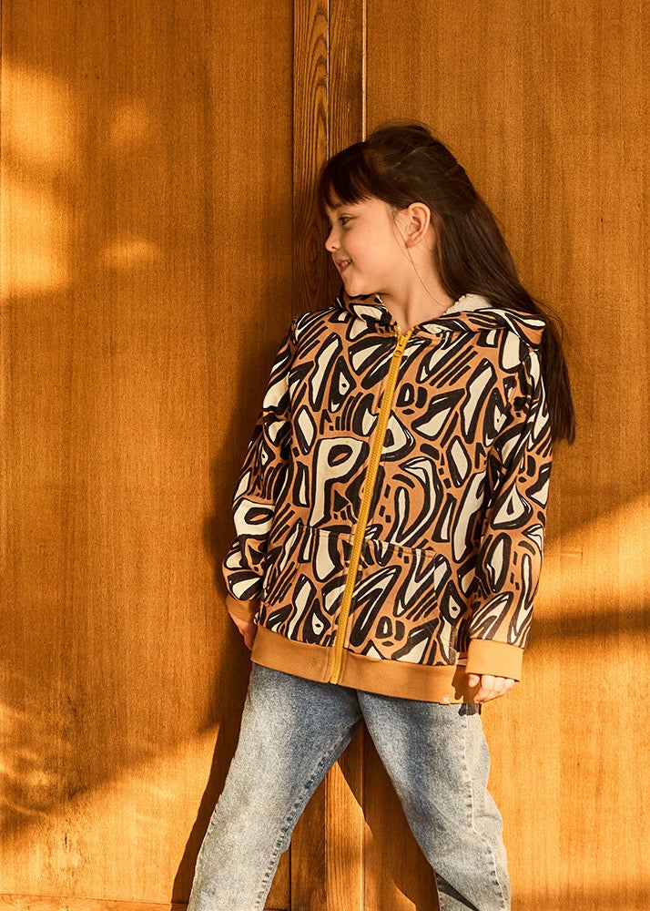 Young girl wearing a patterned jacket standing against a wooden wall.
