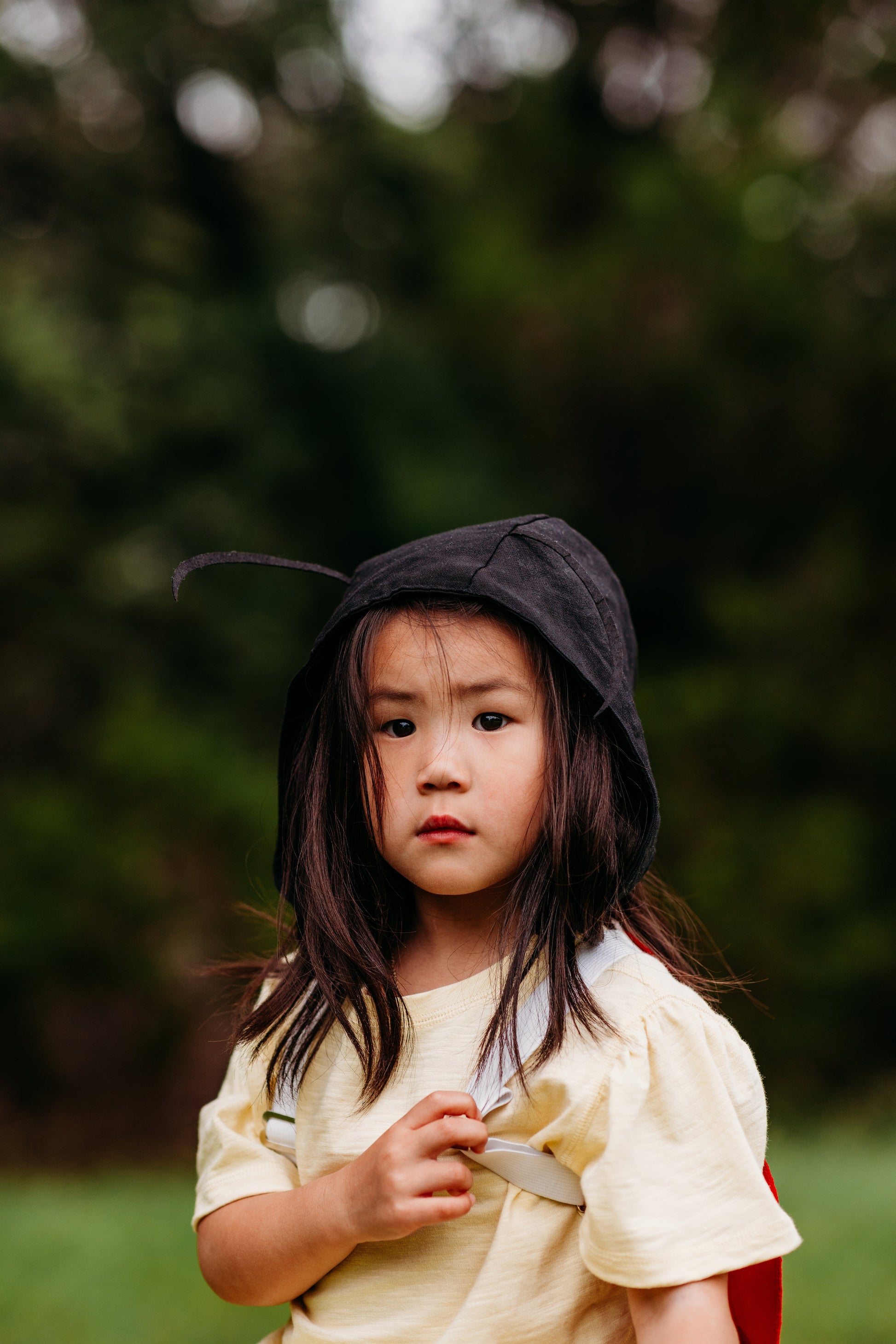 Young girl in a yellow dress with a dark hood standing outdoors with blurred greenery in the background