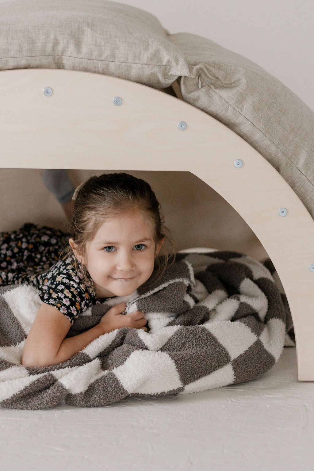 Child lying inside a wooden arch structure with a checkered blanket, surrounded by pillows.