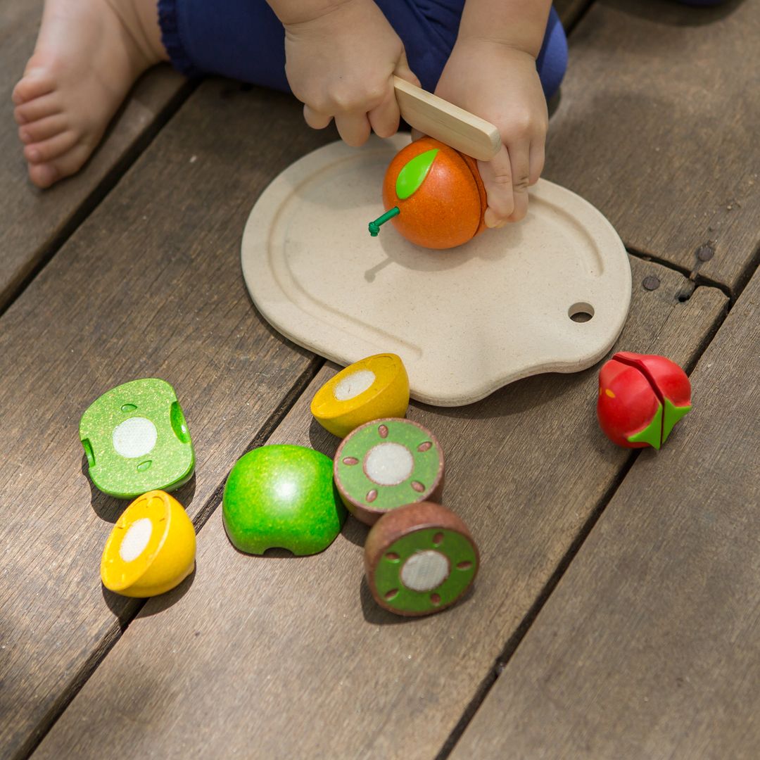 Child playing with wooden toy fruits and a cutting board on a wooden floor.