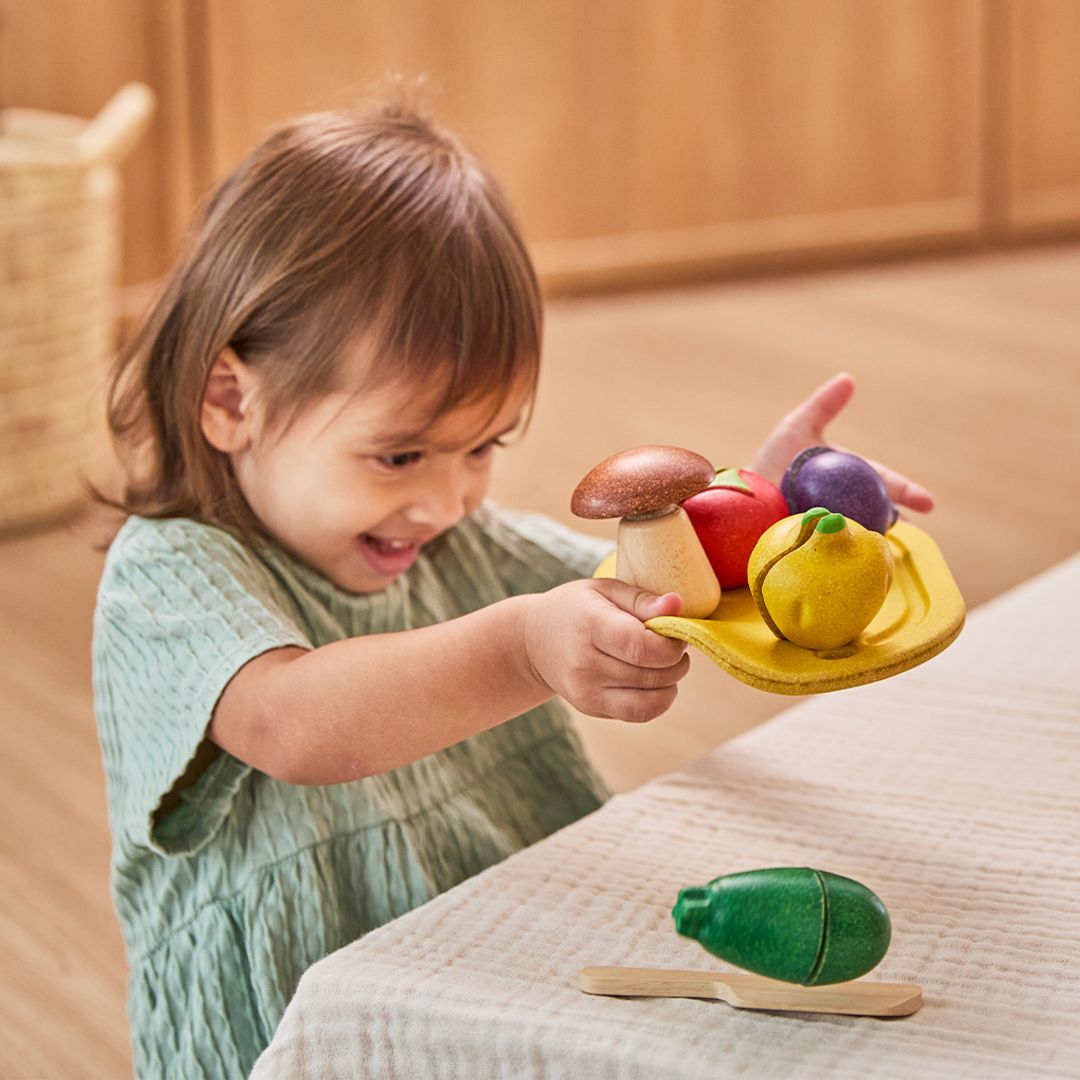 Child playing with colorful wooden toy fruits and vegetables on a light surface.