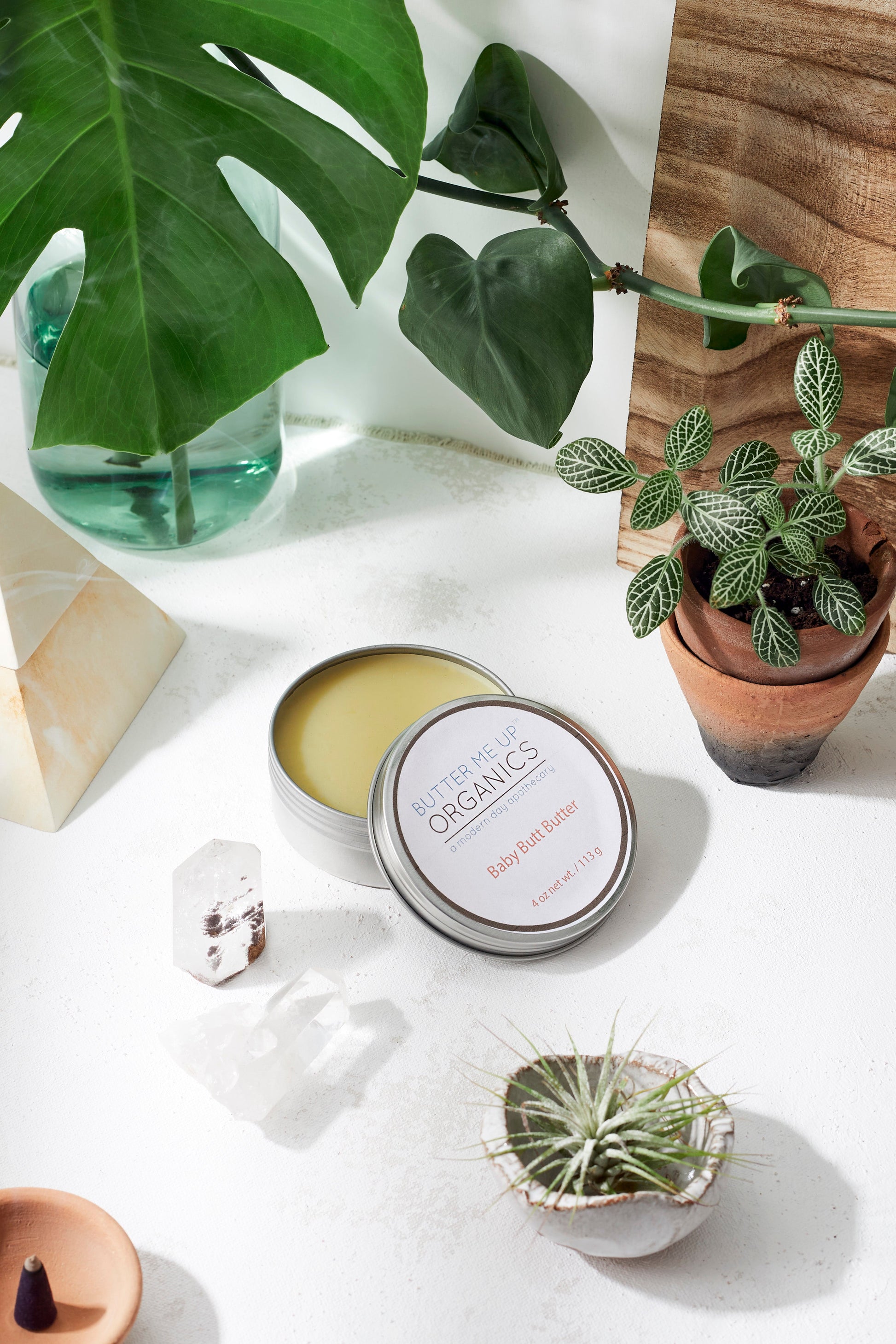 Tin of body butter with a plant and crystal on a white surface