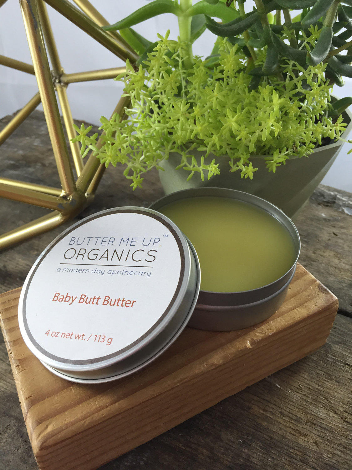 Butter Me Up Organics Baby Butt Butter on a wooden coaster with a plant in the background