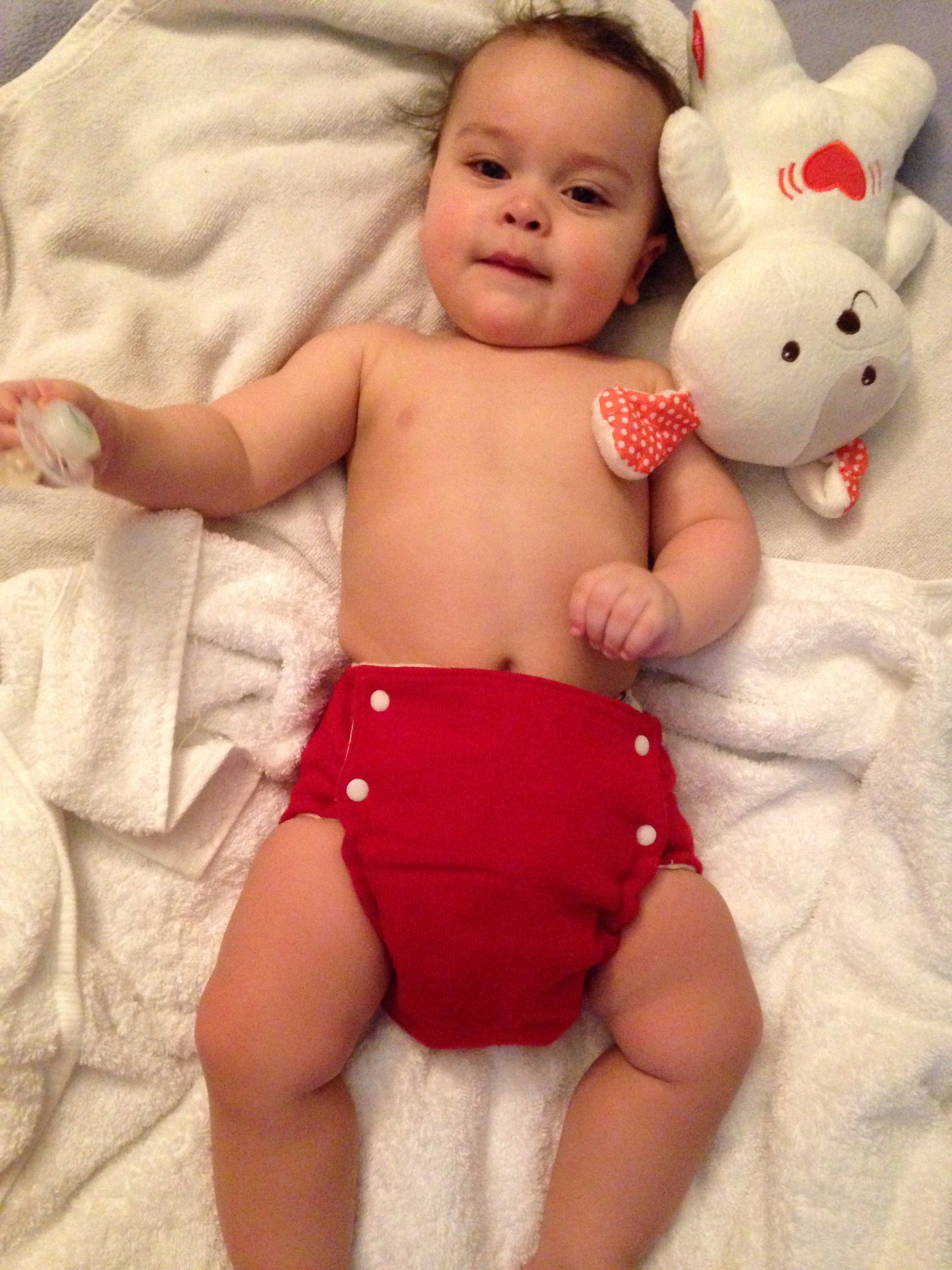 Baby lying on a white blanket wearing a red diaper with white buttons.