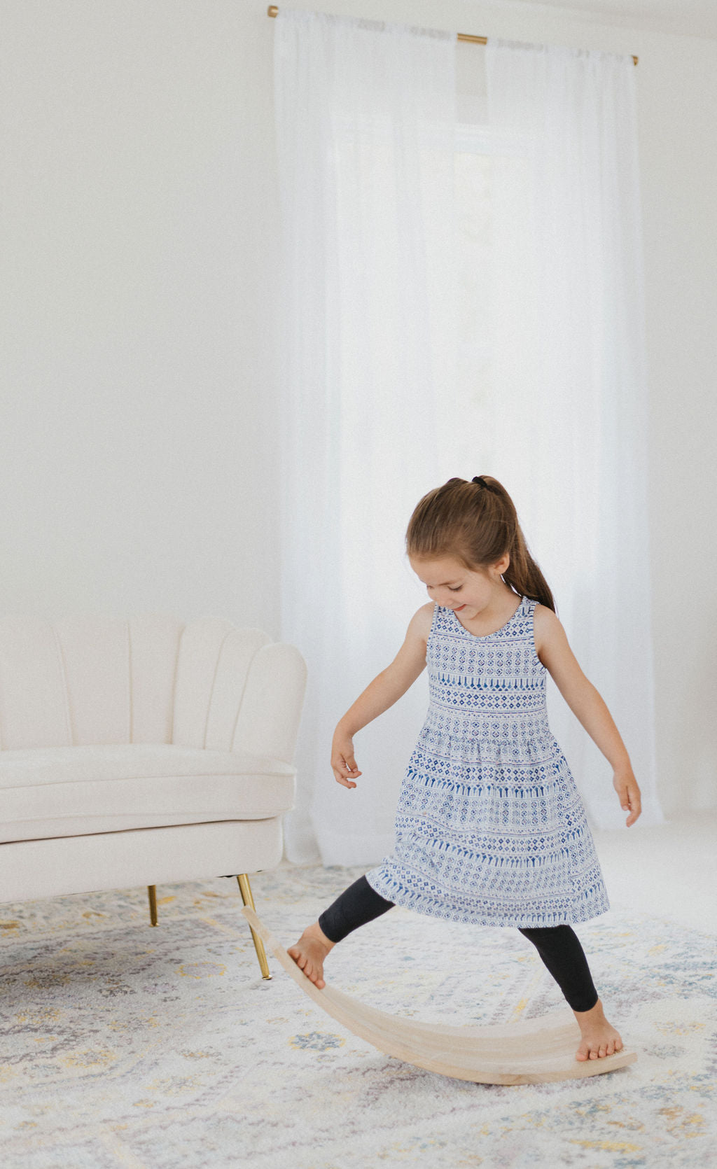 Young girl in a patterned dress standing on a round wooden stool in a room with white curtains and a white sofa.