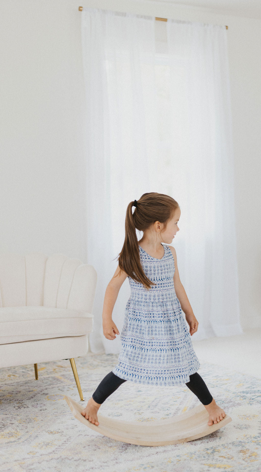 Young girl in a blue dress standing on a wooden stool in a room with white curtains and a white couch.