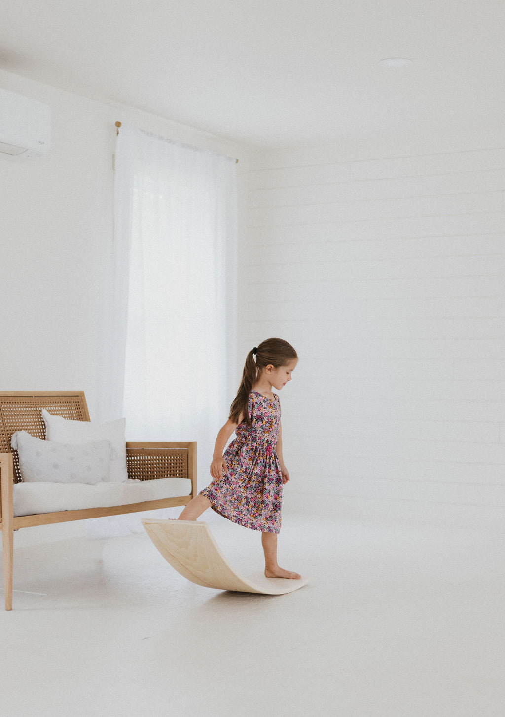 Young girl in a floral dress standing on a wooden stool in a minimalistic room with a white sofa and light-colored floor.