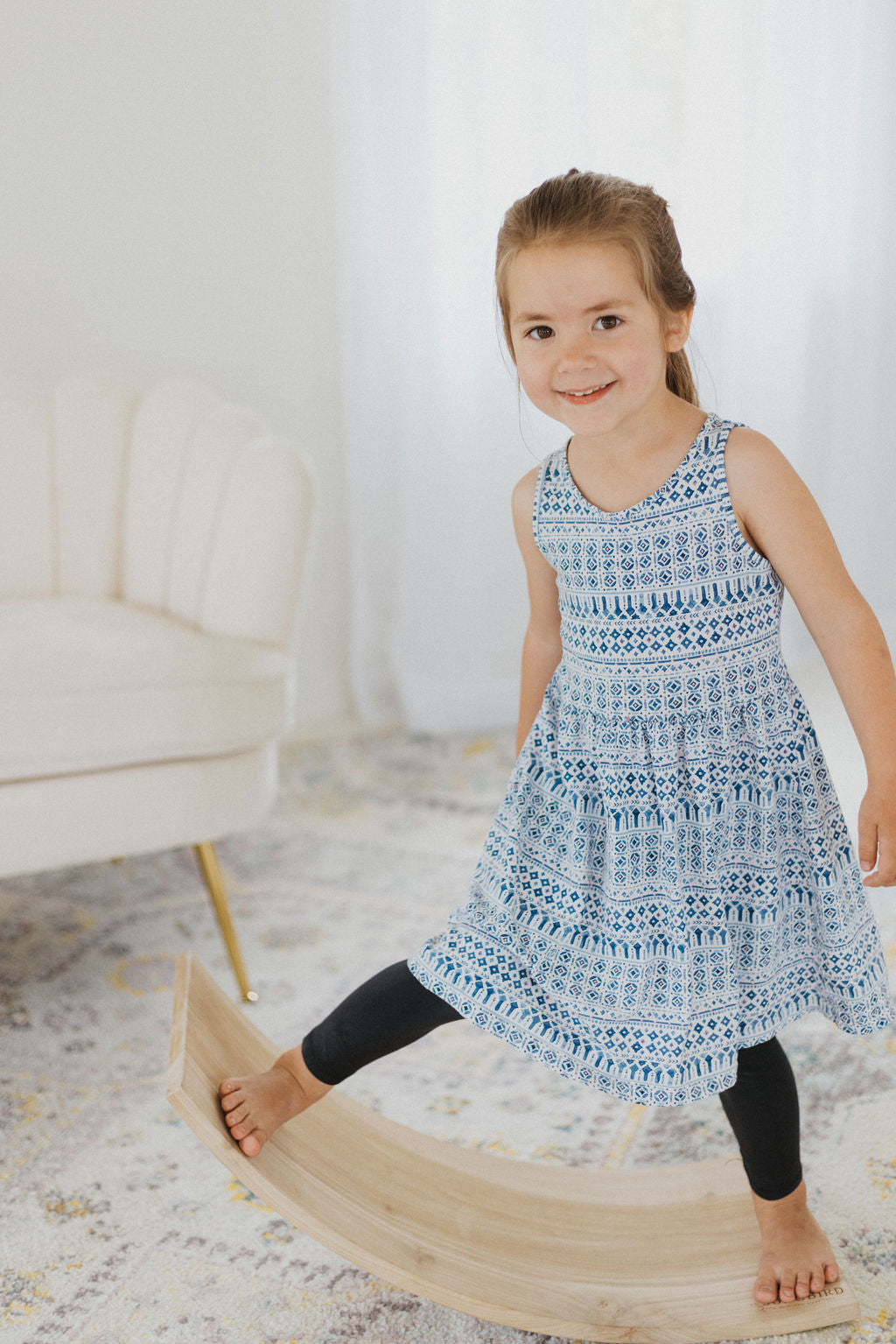 Young girl in a blue patterned dress standing on a wooden balance beam.