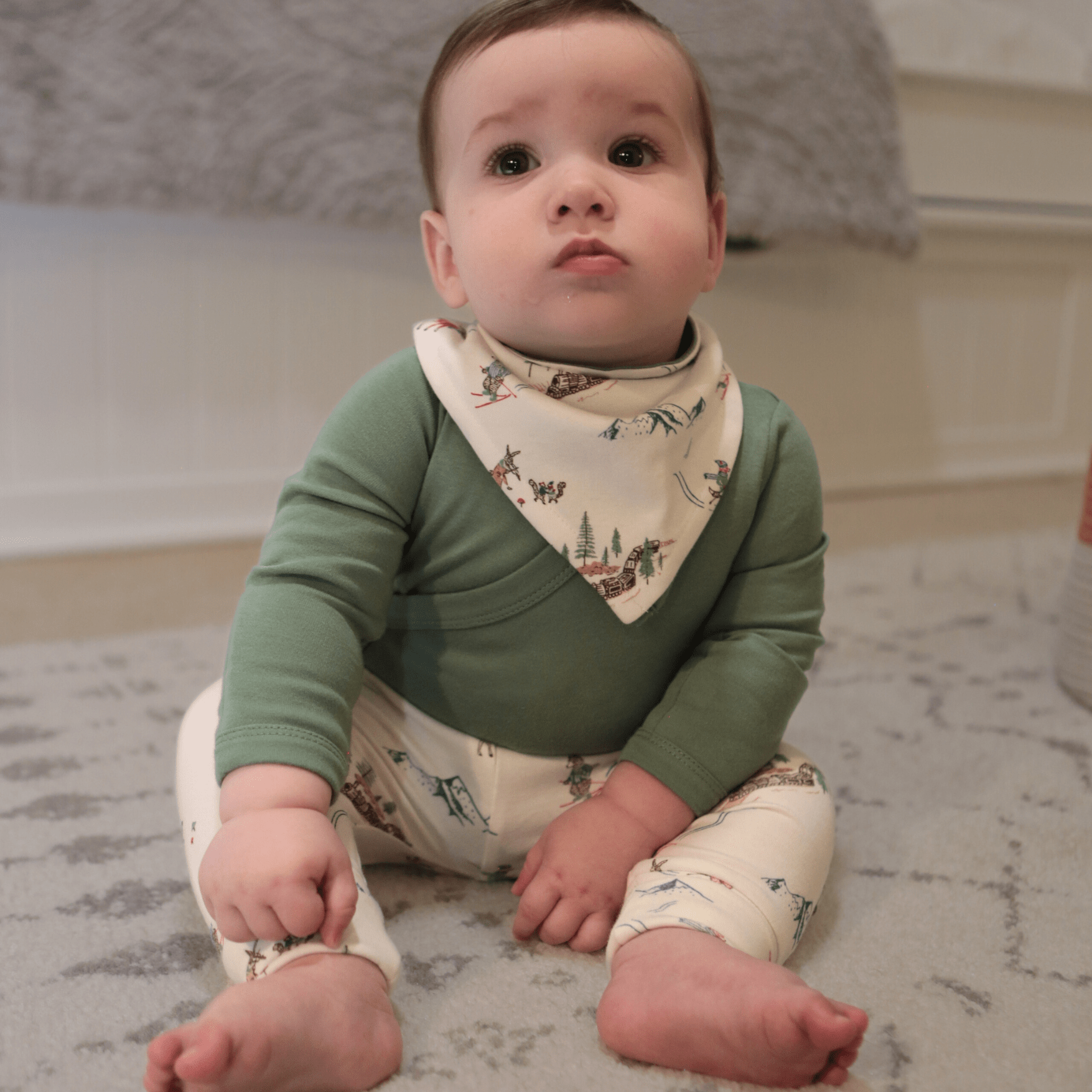 Baby sitting on a carpeted floor wearing a green long-sleeve shirt and patterned pants.
