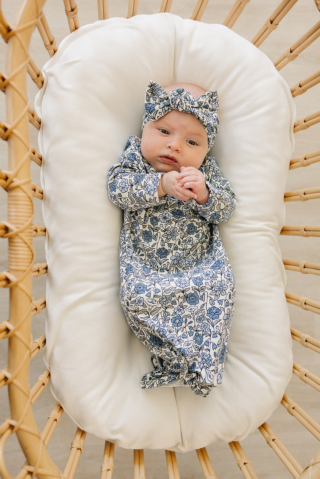 Baby wrapped in a floral swaddle and hat lying on a white cushion in a wicker basket.