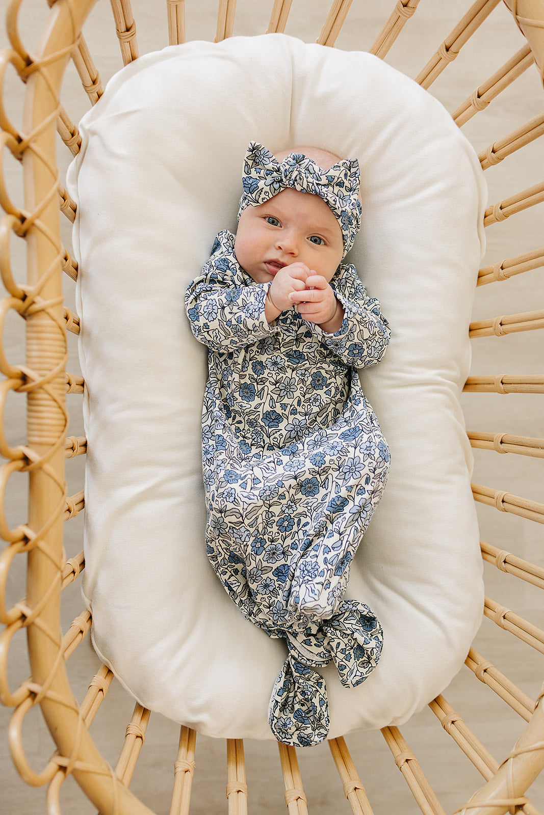 Newborn baby in a floral outfit lying on a white cushion inside a wicker basket.