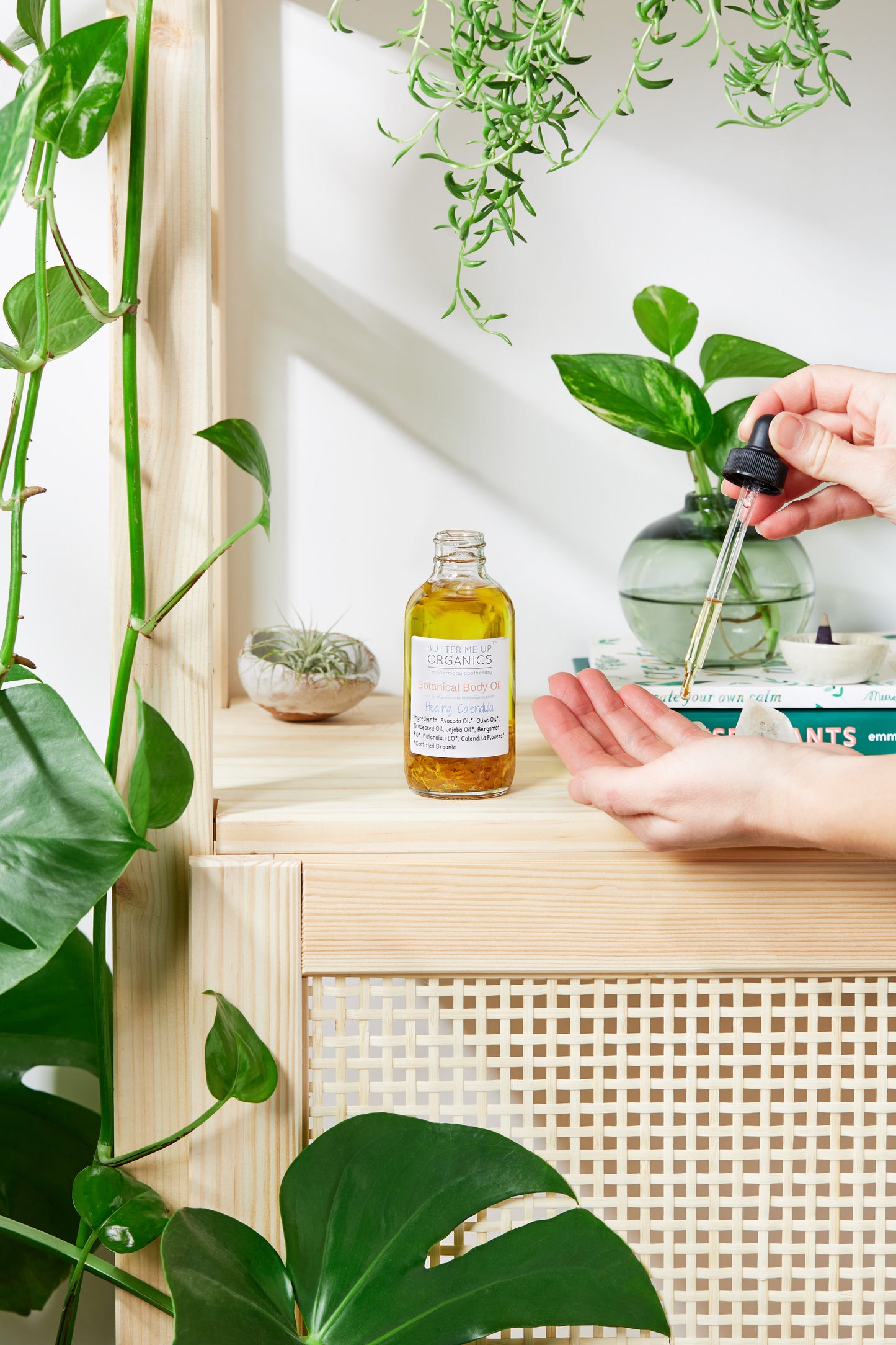 Hand holding a dropper over a bottle of oil on a wooden shelf with plants around