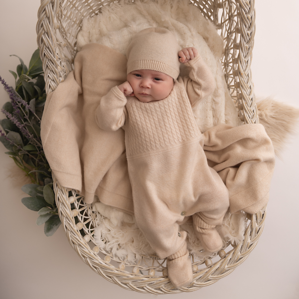 Newborn baby in a beige outfit lying in a wicker basket with soft textures and plants.