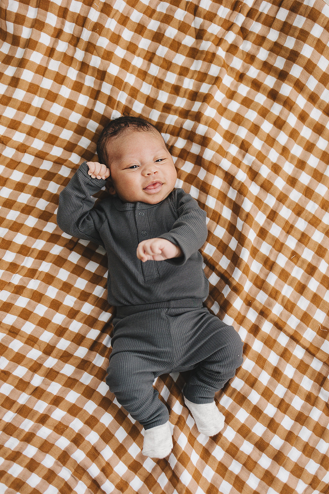 Baby in a gray outfit lying on a brown and white checkered blanket