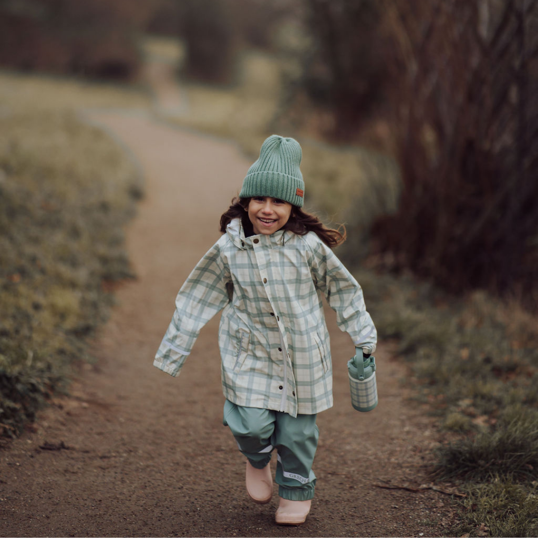 Child in a plaid coat and green hat walking on a path in a natural setting