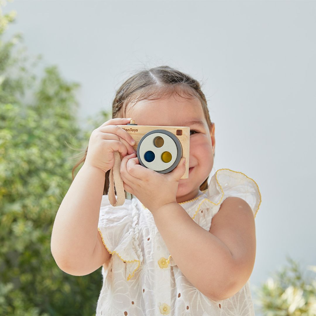 Child holding a toy camera outdoors with greenery in the background