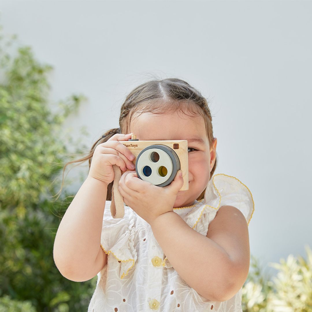 Child holding a toy camera outdoors with greenery in the background