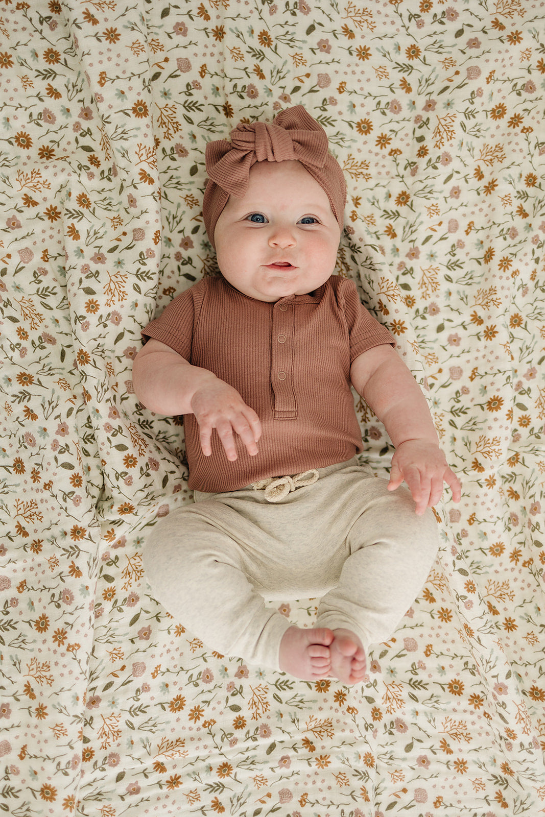 Baby lying on a floral-patterned blanket wearing a brown top and beige pants.