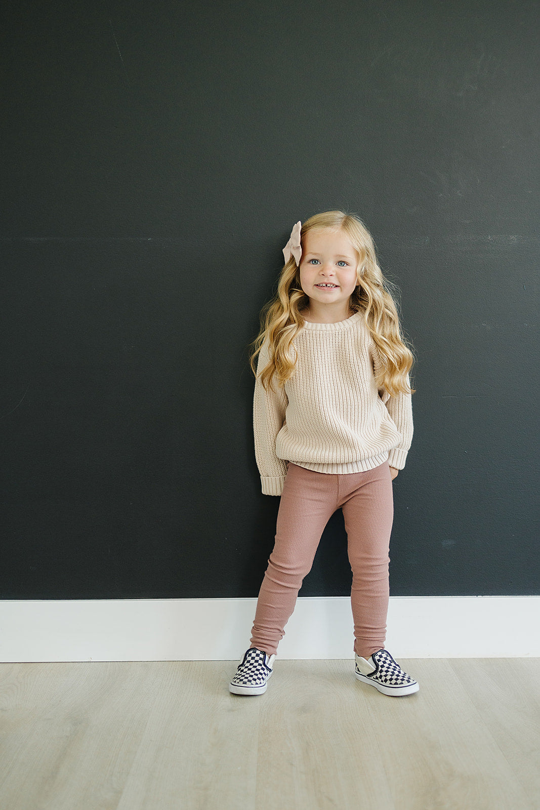 Young girl standing against a black wall wearing a beige sweater and pink pants.