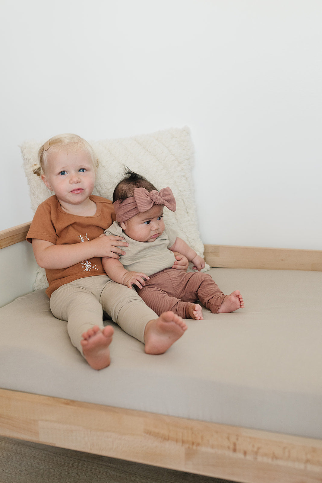 Two children sitting on a bed with a white pillow and wooden headboard.