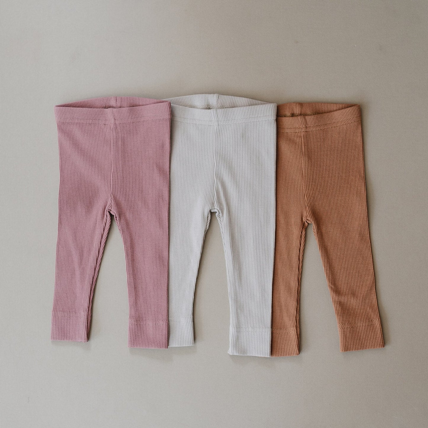 Three pairs of ribbed leggings in pink, white, and brown on a beige background