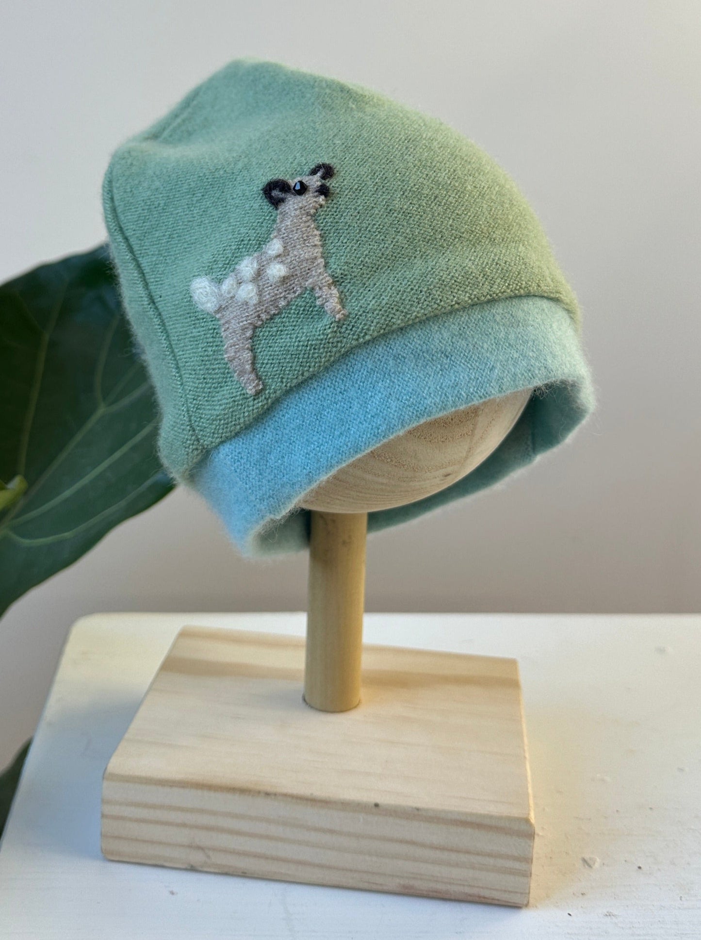 Green and blue baby hat with a embroidered design on a wooden mannequin head.