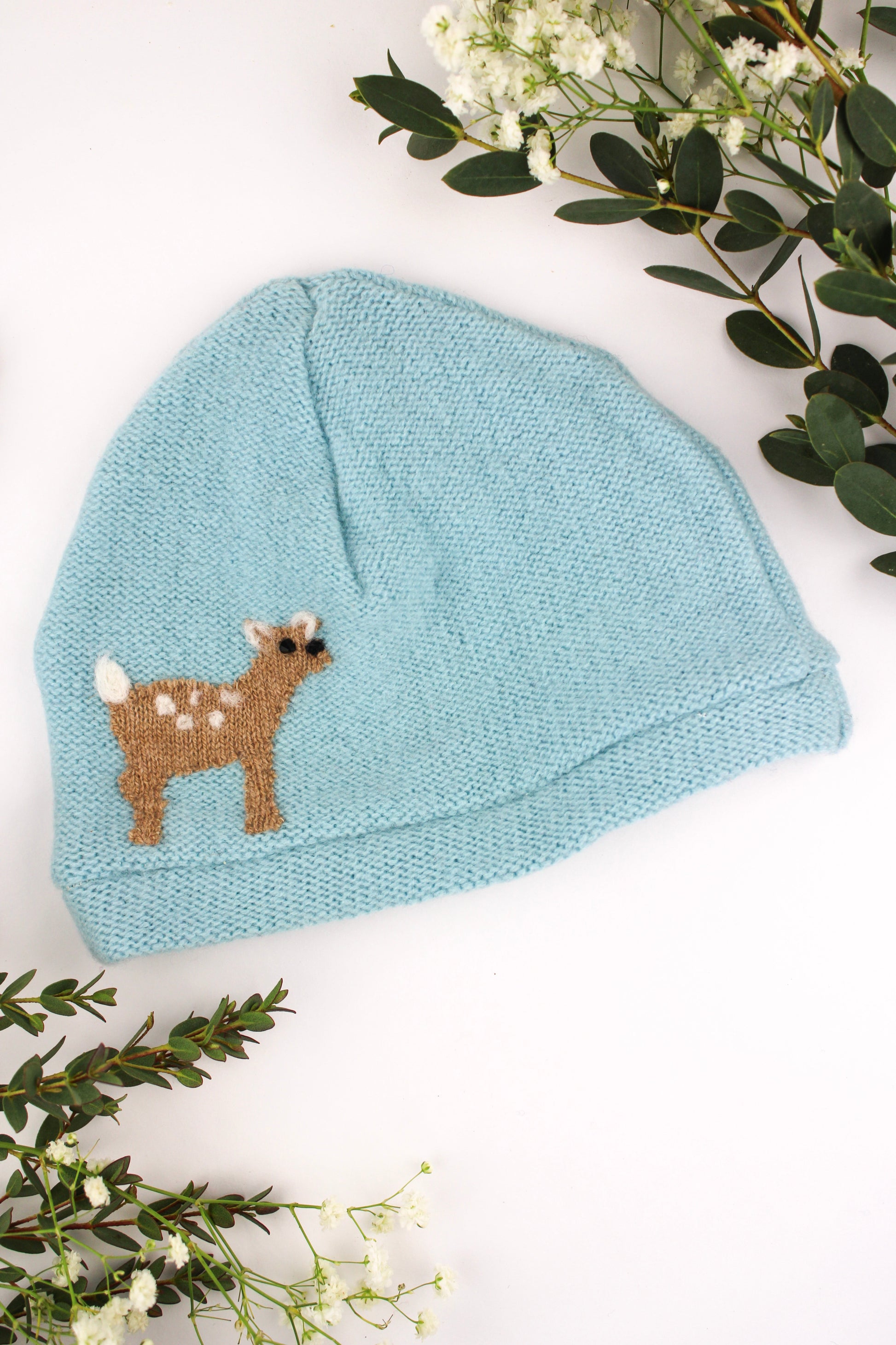 Blue knit baby hat with a deer design on a white background with greenery.