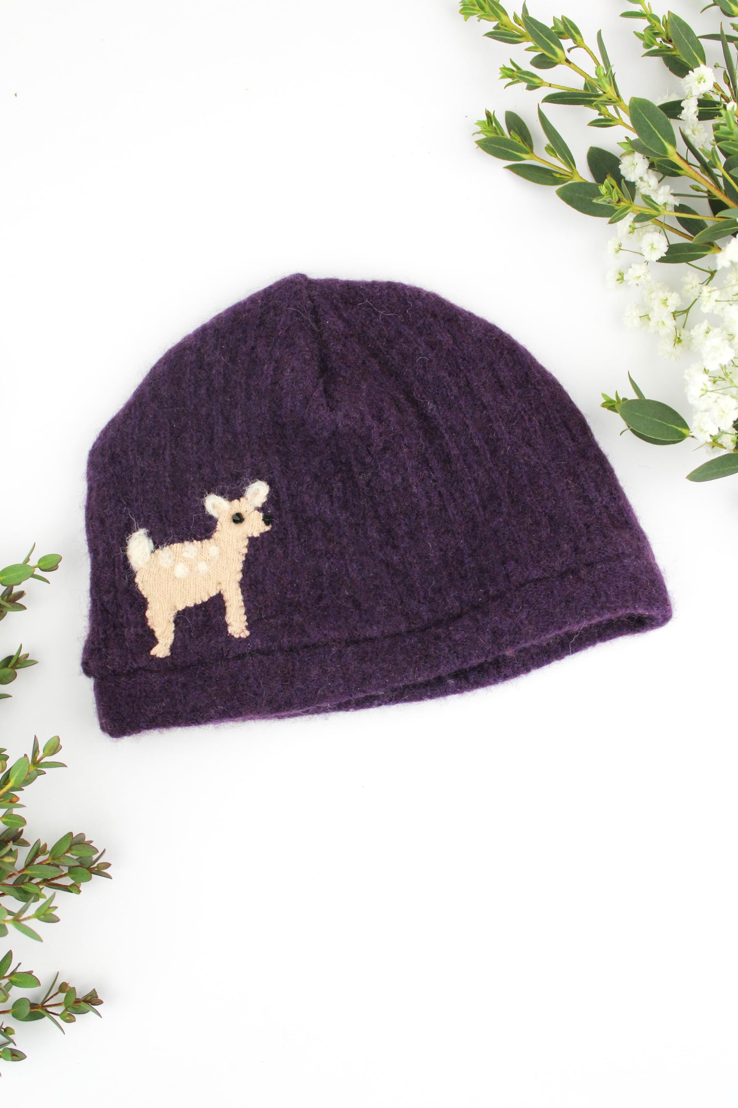Purple knit baby hat with a small dog design on a white background with greenery