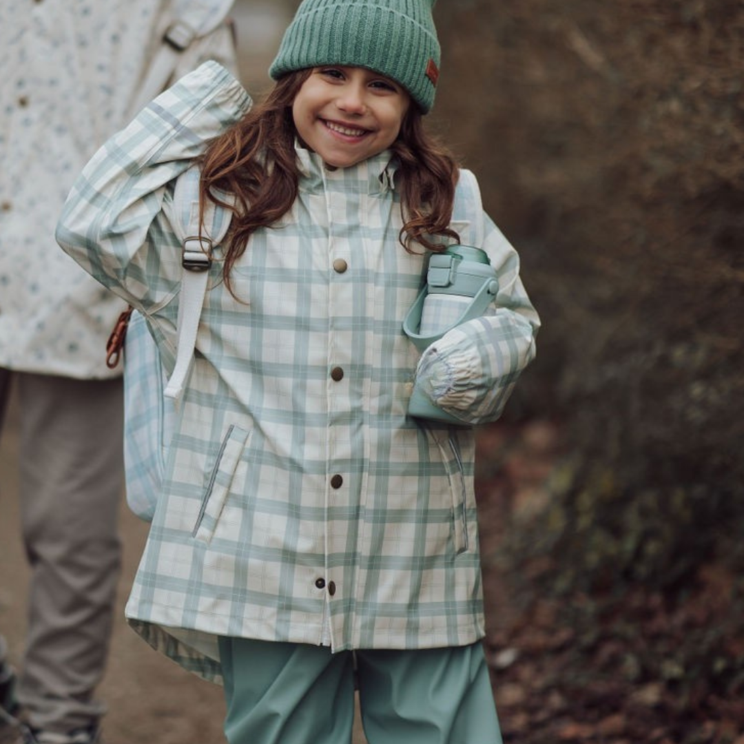 Child wearing a plaid coat and green hat, standing outdoors with a blurred background