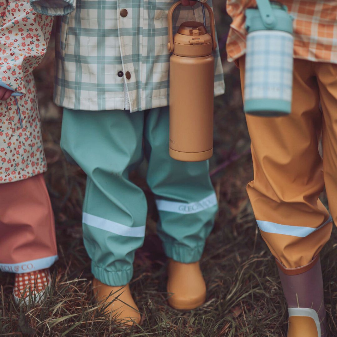 Children wearing colorful raincoats and pants with water bottles in a natural setting.