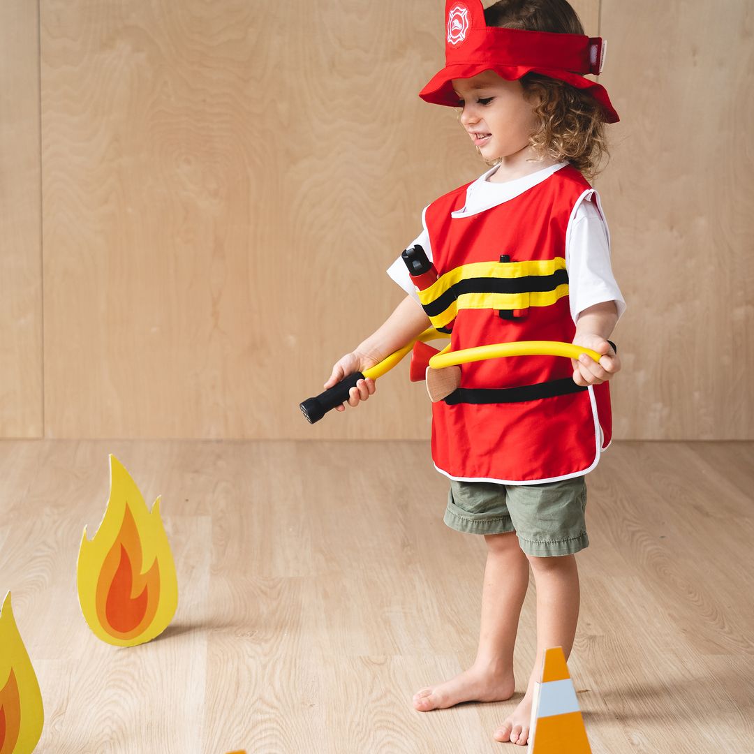 Child in firefighter costume holding a hose on a wooden floor with fire-themed decorations.