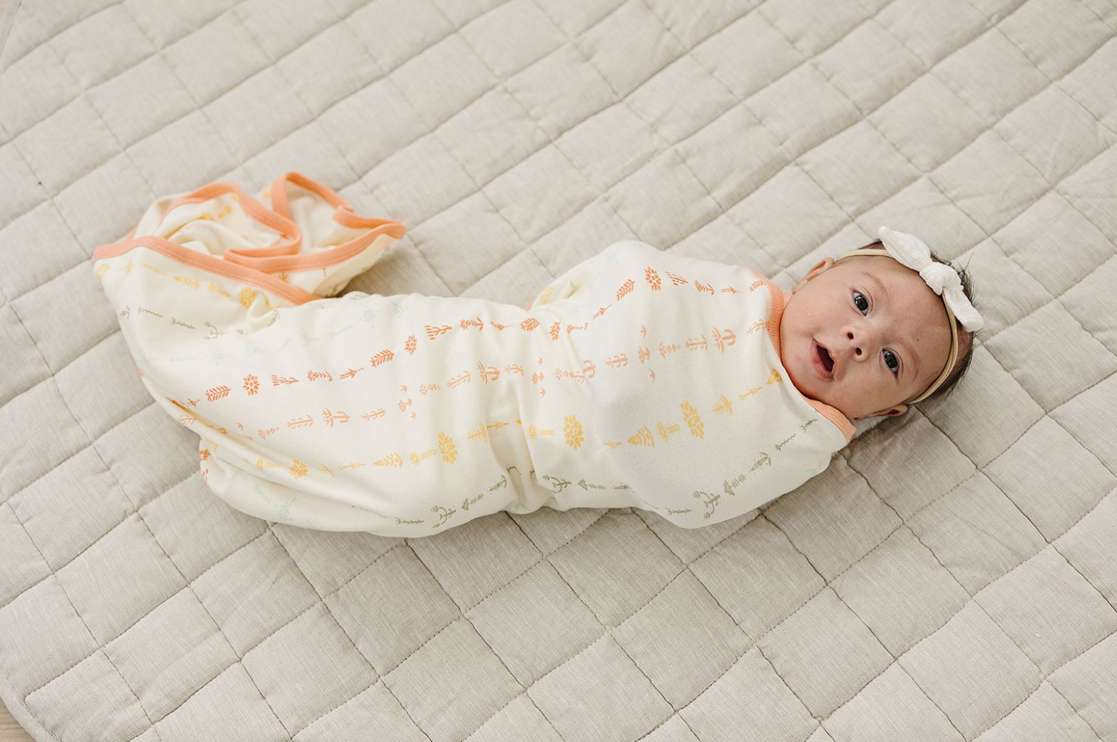 Baby swaddled in a white blanket with orange patterns on a beige quilted surface