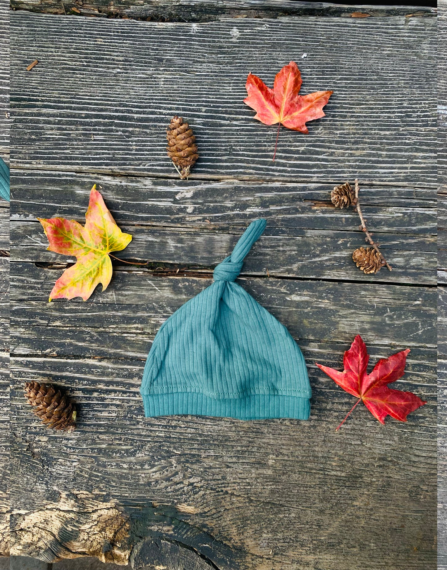 Green ribbed knit baby hat on a wooden surface with autumn leaves and pinecones.