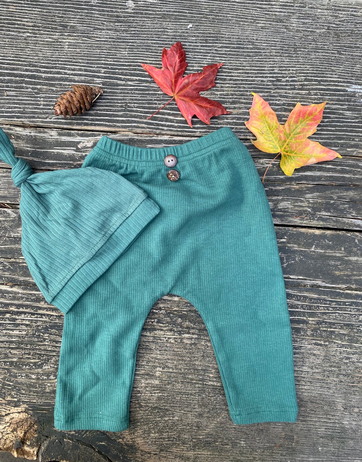Green baby pants and hat  on a wooden surface with autumn leaves and a pine cone.