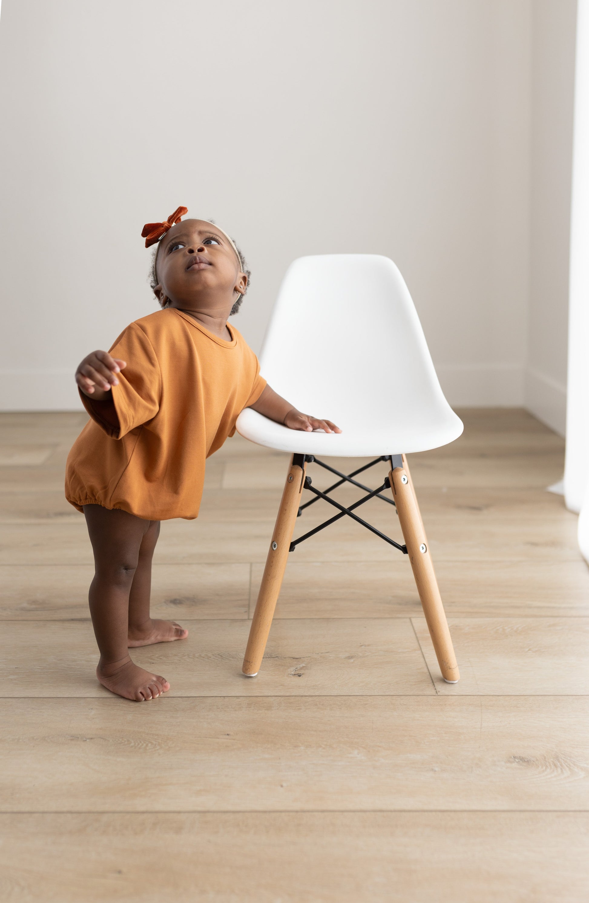 Child in an orange outfit standing next to a white chair with wooden legs on a light wood floor.