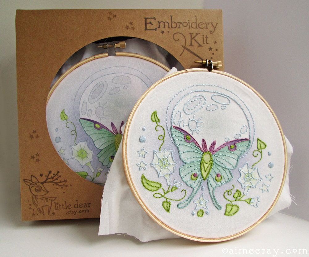 Embroidery kit with a completed butterfly design on fabric inside an embroidery hoop, next to a cardboard box labeled 'Embroidery Kit'.