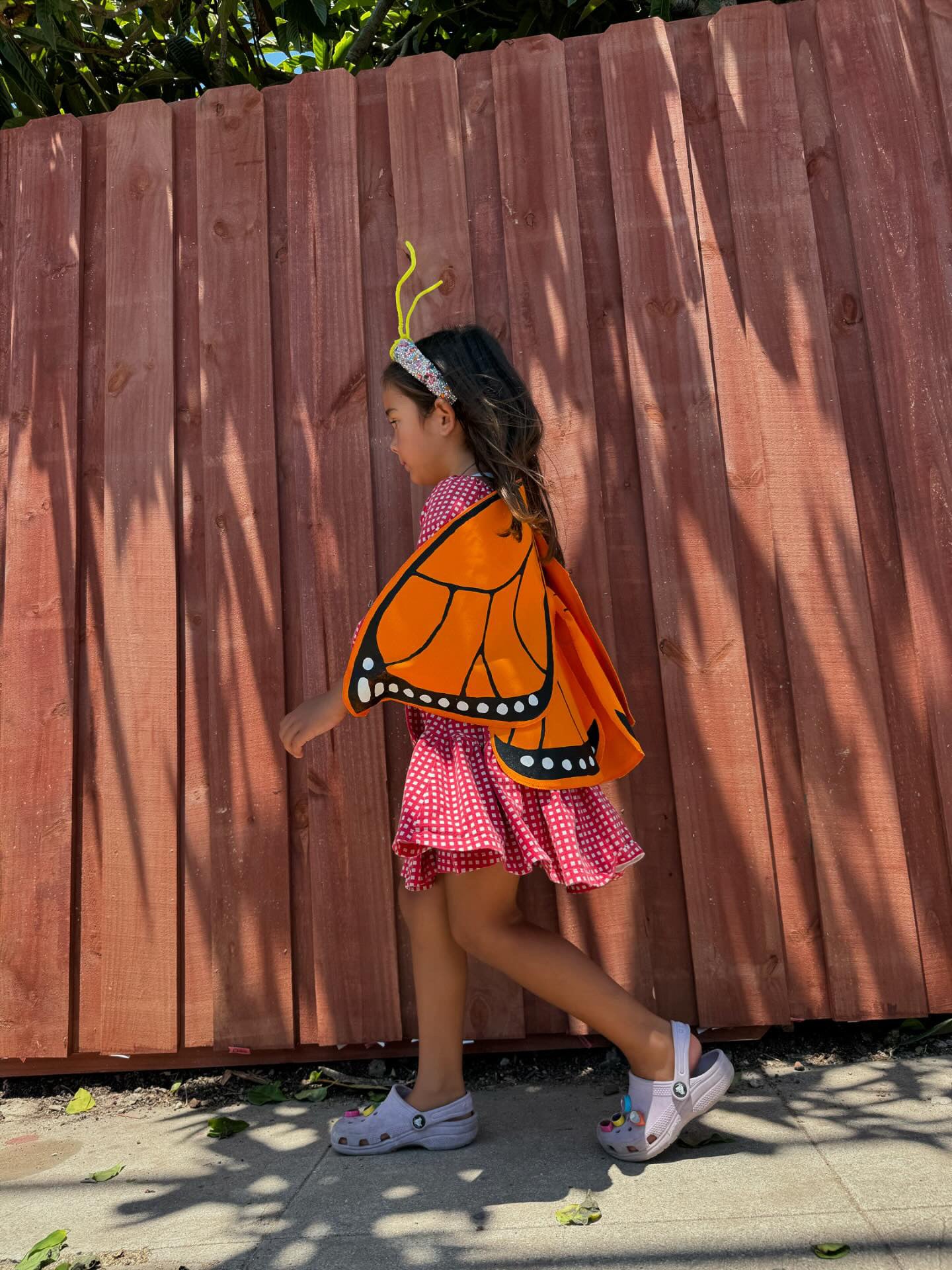 Child wearing a butterfly costume walking in front of a wooden fence.