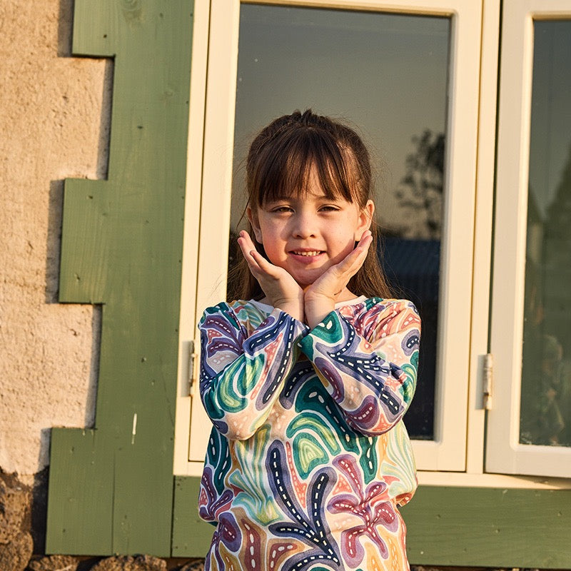 Young girl wearing a colorful patterned shirt standing in front of a window.